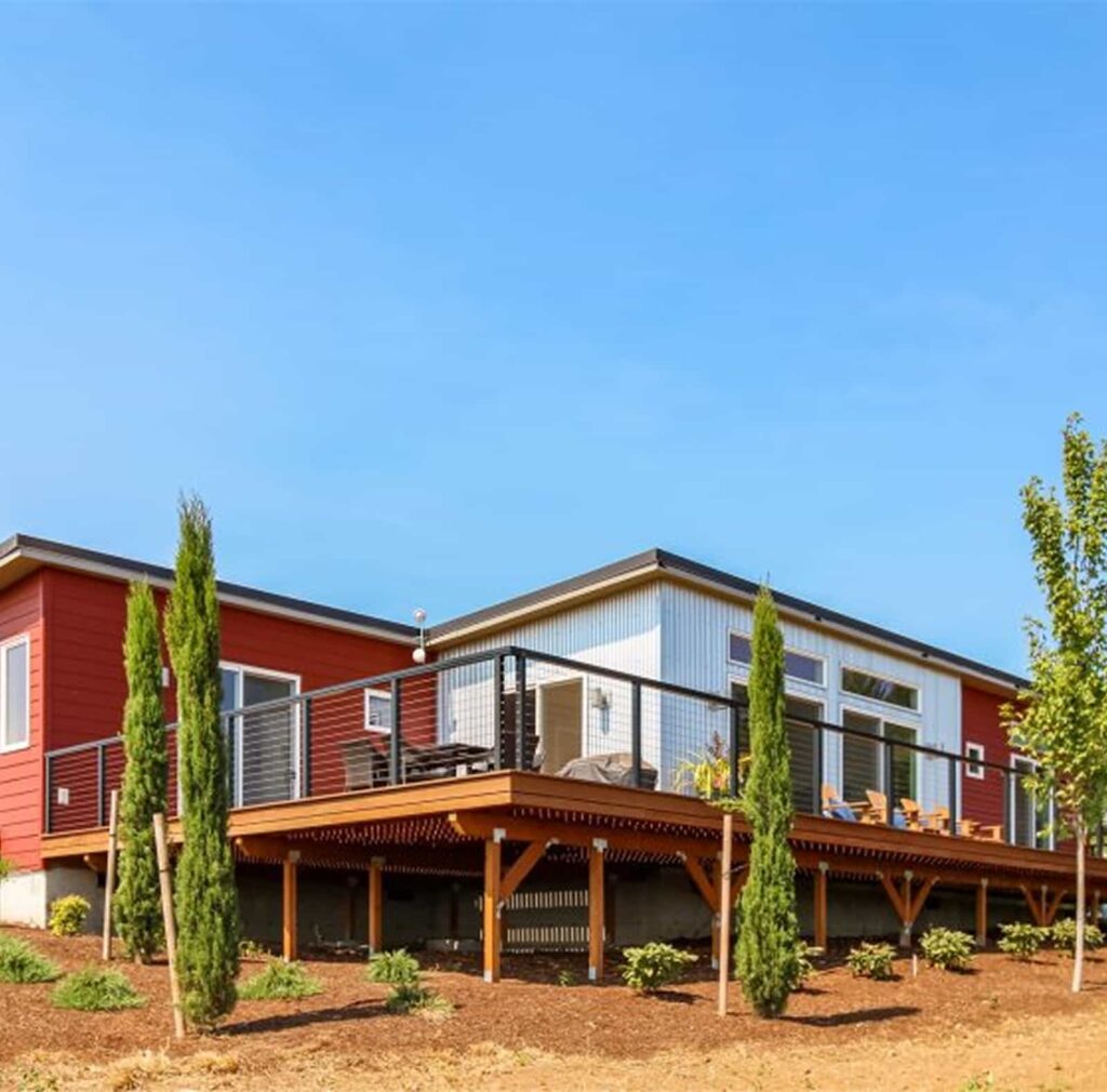 Bryn Mawr prefab home exterior with red and corrugated metal siding, elevated wooden deck with cable railing, and large windows.