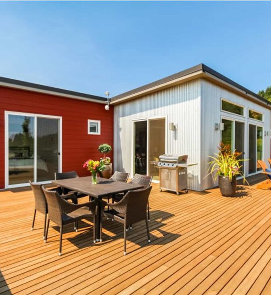 Outdoor deck with wicker dining set, barbecue grill, and sliding glass doors, adjacent to a modern red and white cottage under a clear blue sky.