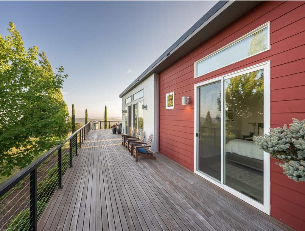 Spacious wooden deck with Adirondack chairs, black cable railing, and large glass doors opening to a red and white modern cottage, surrounded by trees.