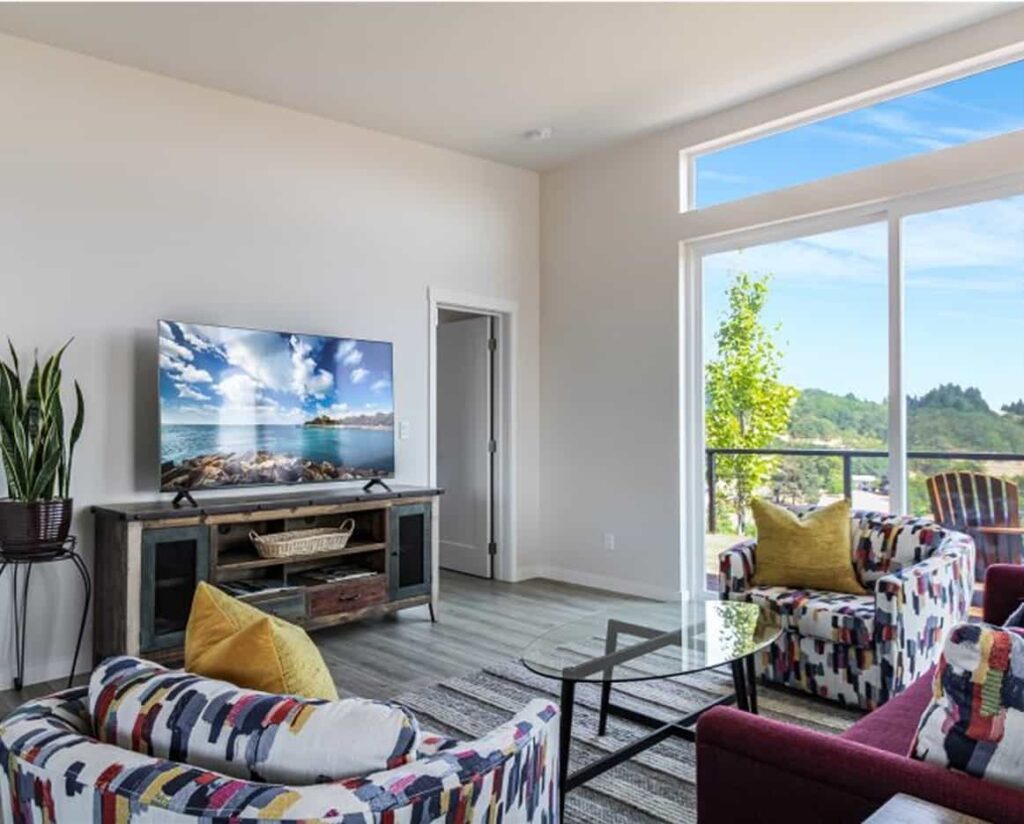 Modern living room with colorful patterned armchairs, a large flat-screen TV on a rustic console, and floor-to-ceiling windows showing lush green hills.