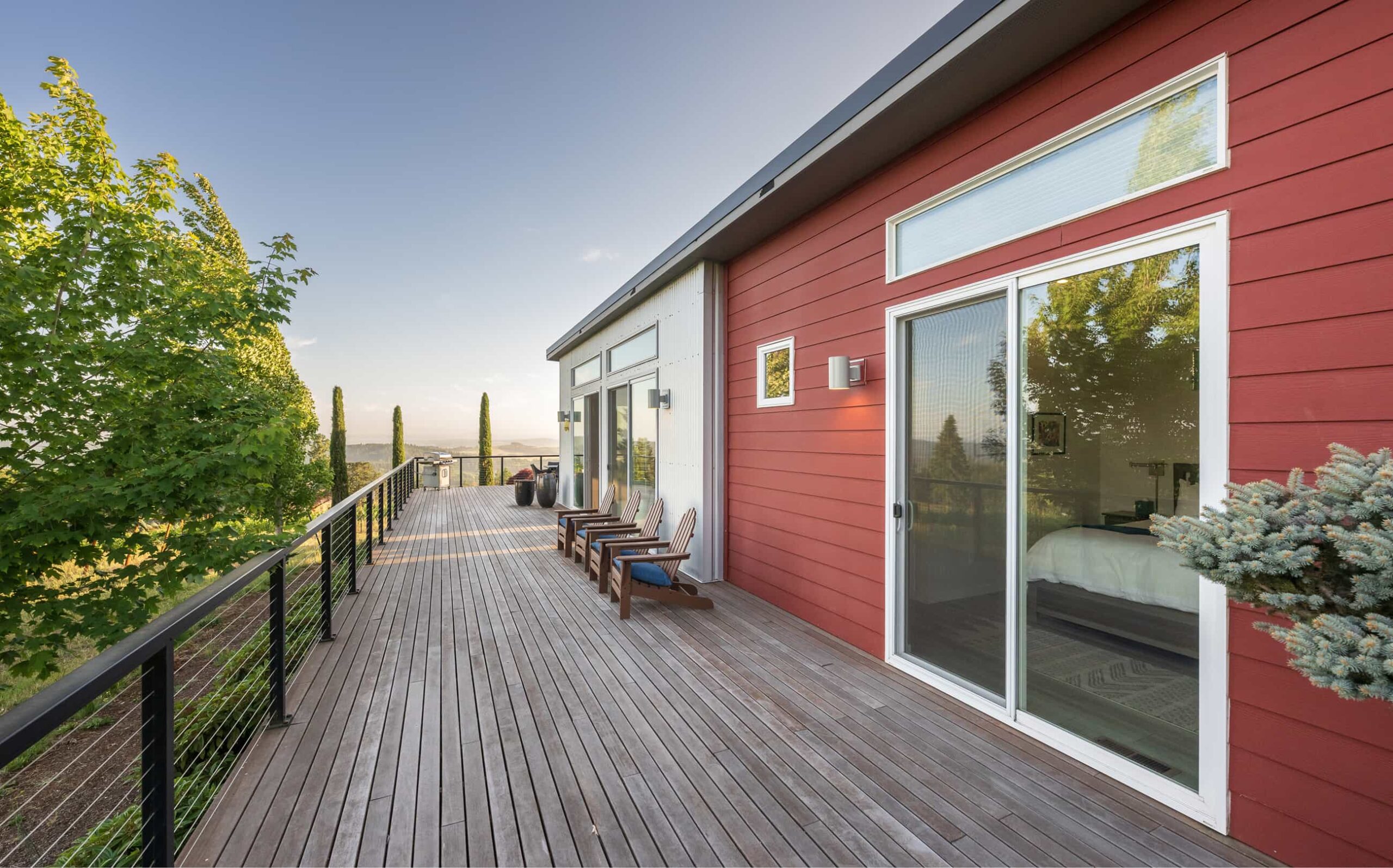 Elevated modern home deck with red siding, lounge chairs, and scenic tree-lined hillside view