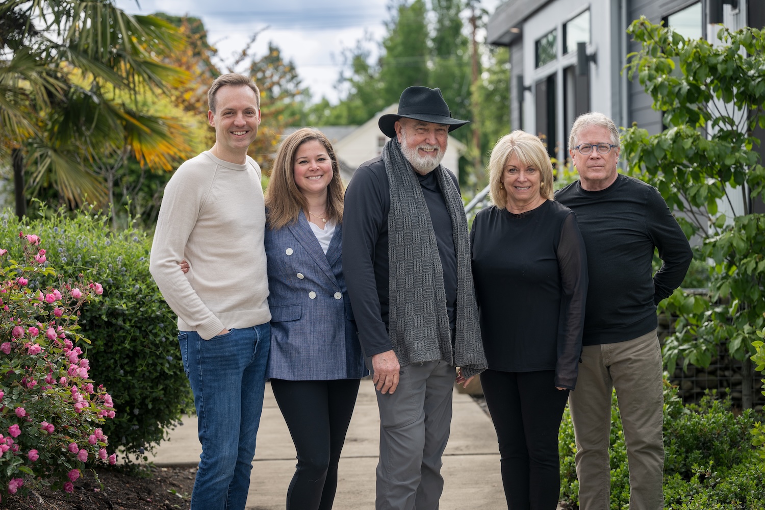 Five members of the IdeaBox team smiling outdoors on a garden path beside blooming shrubs, standing casually in front of a modern model home.