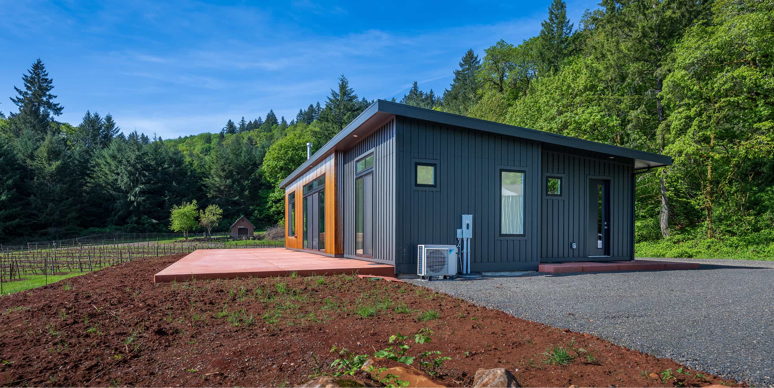 Modern dark gray guest cottage with vertical siding and natural wood accents, surrounded by forest and vineyard, under a clear blue sky.