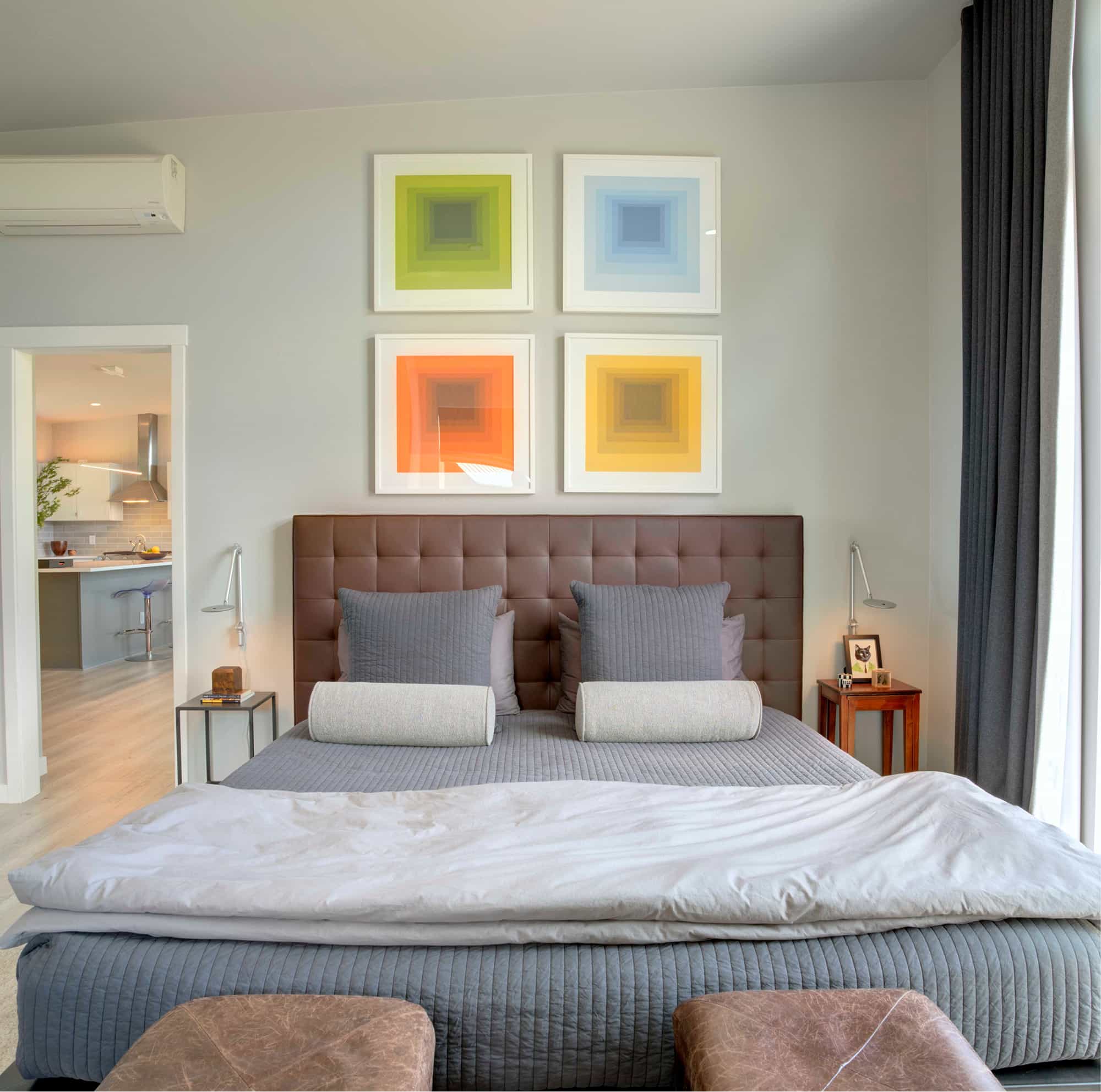 A modern bedroom interior featuring a tufted headboard, gray bedding, and four colorful square gradient artworks above the bed, with a view into the kitchen area.