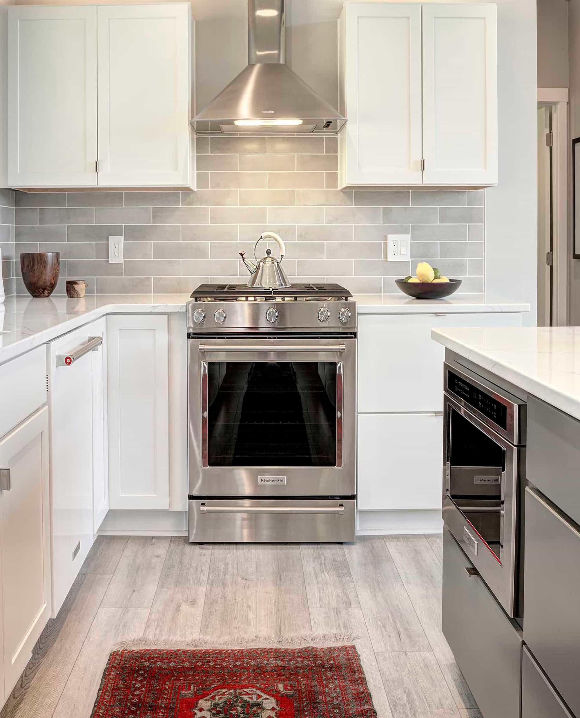 A contemporary kitchen with white cabinets, gray subway tile backsplash, stainless steel appliances, and light wood flooring, accented by a red patterned rug.