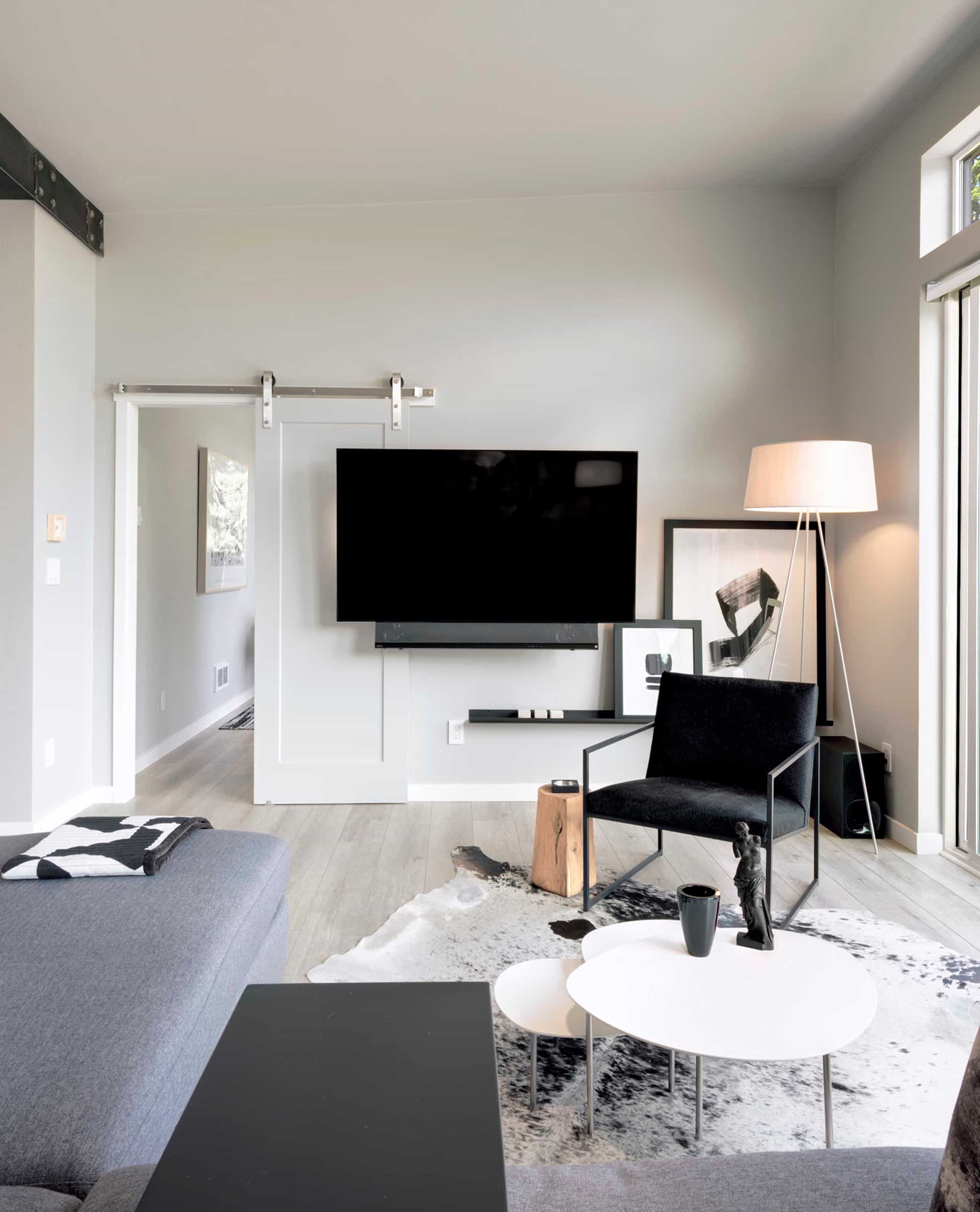 A stylish living room with minimalist furniture including a black chair, nested white coffee tables, and a gray sectional, centered around a large TV mounted on the wall and decorated with modern art and a floor lamp.