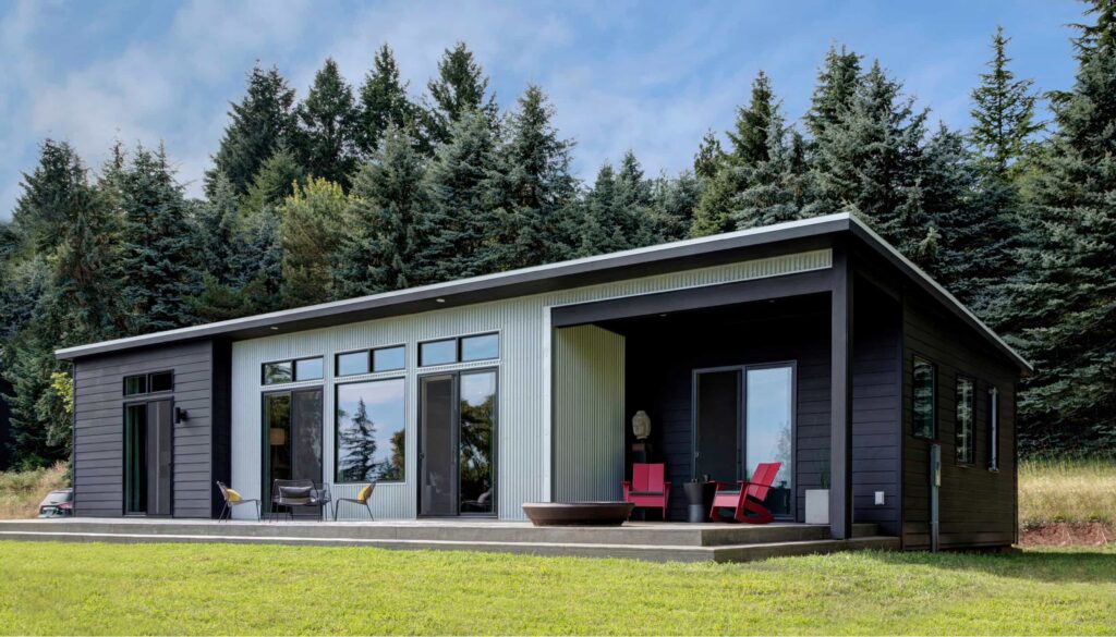 Modern black and white modular home with large windows, red Adirondack chairs, and forest backdrop on a grassy lot